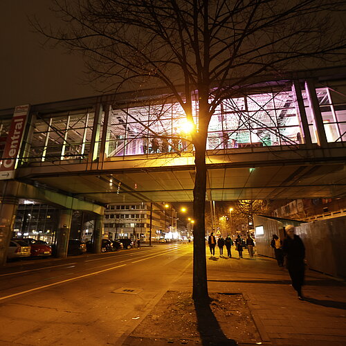 Außenansicht der Brücke über die Worringer Straße bei Nacht. Die Brücke ist von innen beleuchtet, Leute bevölkern die Brücke.