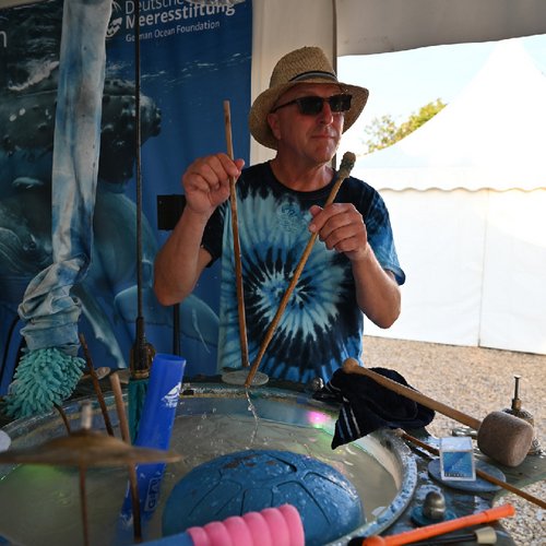 Foto des Musikers Konrad Zimmermann mit einem Batiktshirt und Hut, der Musik macht. Auf dem Tisch vor ihm sind verschiedene Musikinstrumente zu sehen.